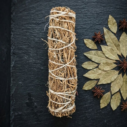 A natural dried herb smudge bundle wrapped in twine, displayed on a dark slate board next to dried bay leaves and star anise.
